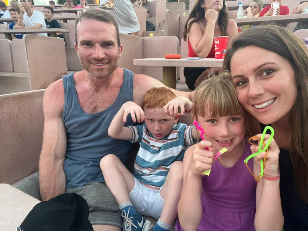 Family holiday photo from Spain, 2024 — the children enjoying the pool while John, visible in the background, looks distant and not himself