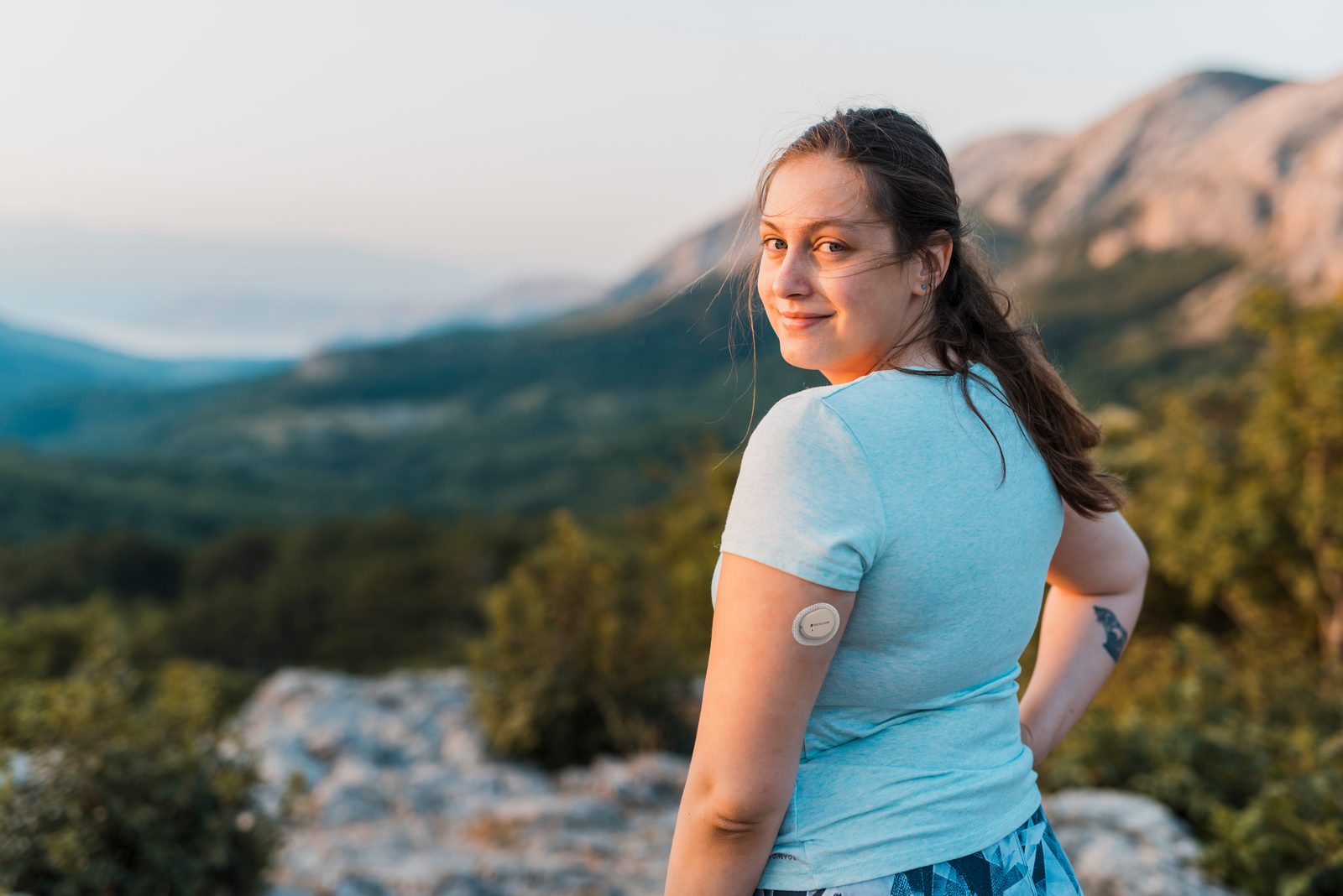 An adult wearing a Dexcom G7 sensor on the back of her arm, looking at a mountain landscape at golden hour